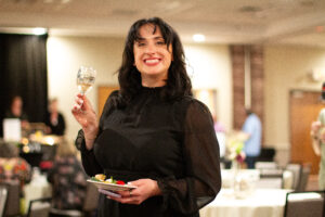 A woman in black formalwear smiling and holding a wine glass and a plate of salad.